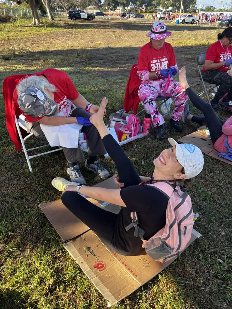 Jennie with her team at a Komen 3-Day. She is sitting on the ground while another teammate checks out her feet from so much walking. She is smiling back at the camera with a white baseball hat on