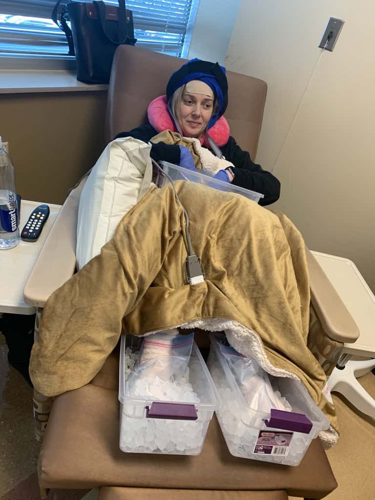 Jennie experiencing cancer treatment with her feet each in a container of ice, covered in a large heated blanket with a hood on