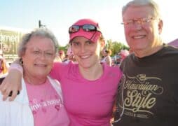 A group of three people, two elderly individuals and a younger person in the middle, pose together, smiling at an outdoor event. They wear pink attire related to the Susan G. Komen 3-Day. A sign in the background reads "Susan G. Komen 3-Day."