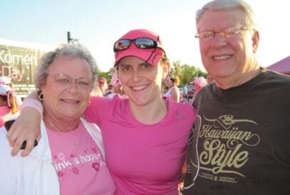 A group of three people, two elderly individuals and a younger person in the middle, pose together, smiling at an outdoor event. They wear pink attire related to the Susan G. Komen 3-Day. A sign in the background reads "Susan G. Komen 3-Day."