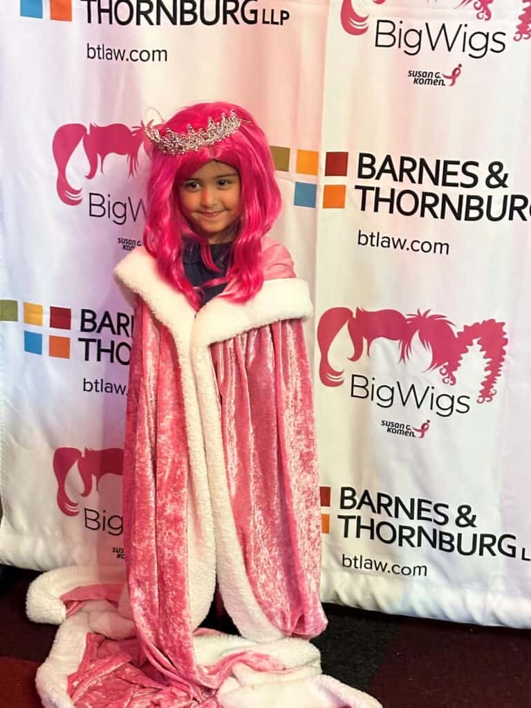 A young girl wearing a hot pink wig, pink robe and silver tiara stands in front of a step-and-repeat banner at a VIPink breast cancer fundraising event. She is smiling and looking away from the camera.