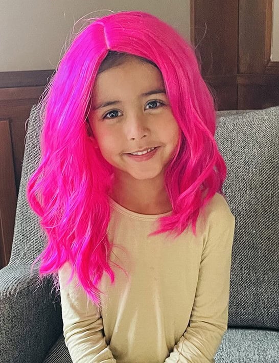 A smiling, young girl wearing a hot pink wig sits on a gray couch. She is wearing a long-sleeved tan shirt.