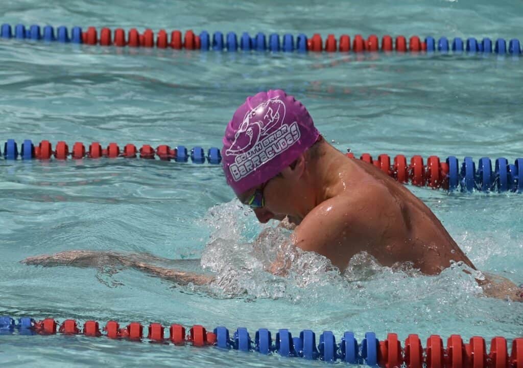 A swimmer wearing a pink swimming cap and goggles competes in a race, performing the breaststroke. The pool has red and blue lane dividers. The swimmer is mid-stroke, with water splashing around them. This event proudly supports "Go pink for the cure" to raise breast cancer awareness.
