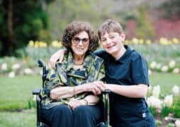 An elderly woman in sunglasses sits in a wheelchair in a garden, smiling, with a young boy beside her, also smiling. The boy has his arm around her shoulder as they pose together. Flowers and greenery form the backdrop.