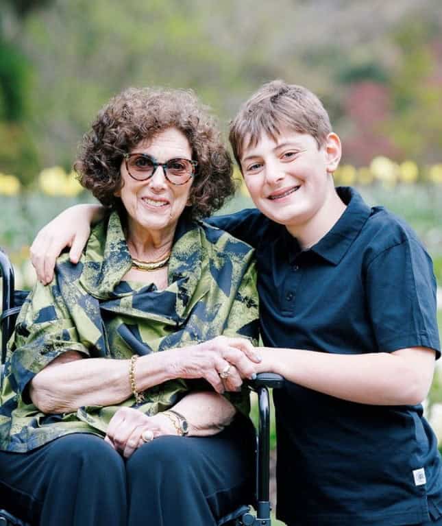 An elderly woman in sunglasses sits in a wheelchair in a garden, smiling, with a young boy beside her, also smiling. The boy has his arm around her shoulder as they pose together. Flowers and greenery form the backdrop.
