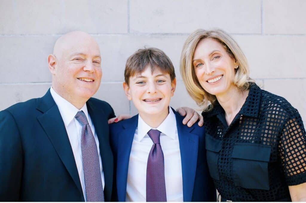 A smiling family of three is posed in front of a light-colored wall. A man in a suit stands on the left, a boy wearing a suit and tie is in the center, and a woman in a black dress is on the right, with her arm around the boy's shoulder.
