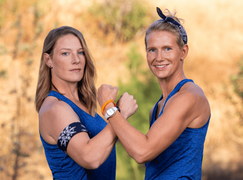 Two women stand outdoors, both with determined expressions and wearing sleeveless blue tops. They face the camera, flexing their biceps and showing off their fitness trackers. One woman wears a headband and the other has a bandana armband. The background is blurred.