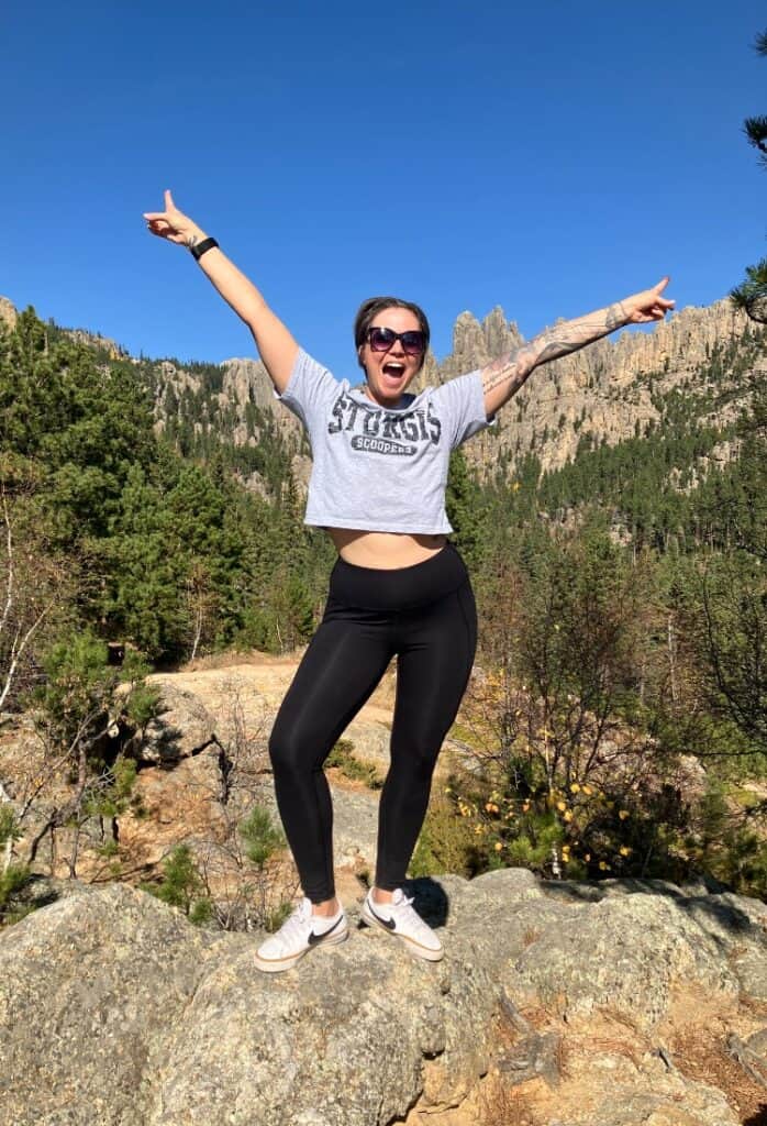 Casey stands in the mountains, arms thrown wide open, a joyful smile on her face.