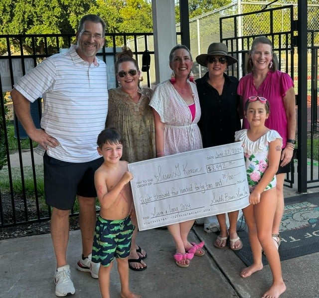 A group of seven people, including two children in swimwear, smiling and holding a large novelty check outdoors by a pool. The check is made out to "Susan G. Komen" for eight thousand thirty-two dollars, supporting the Go Pink for the Cure campaign. They stand on a concrete surface near a fence.
