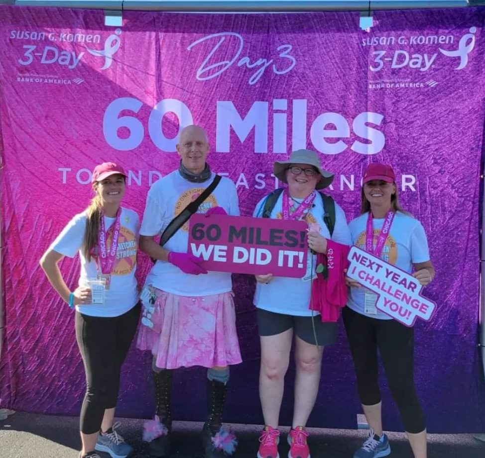 Four people are posing in front of a banner that reads "Susan G. Komen 3-Day, 60 Miles Toward a World Without Breast Cancer." They are celebrating completing a 60-mile walk. Two hold signs that say "60 Miles! We Did It!" and "Next Year I Challenge You!.
