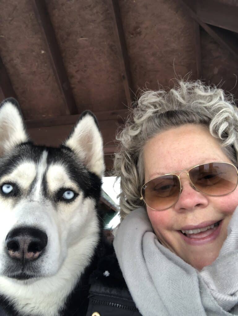 Eve, who had to advocate for herself, smiles, wearing sunglasses, her hair curly. A beautiful black and white husky with bright blue eyes is next to her.