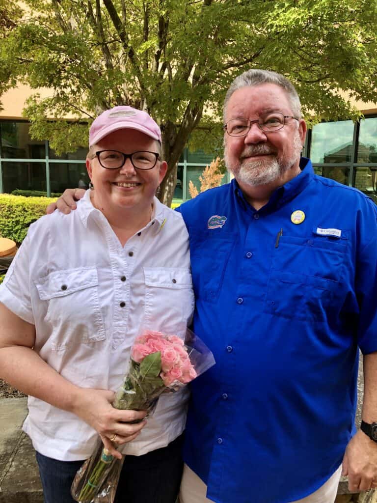 A woman in a pink hat, celebrating her last chemotherapy session, holds pink roses and stands next to her husband, who wears a blue shirt. Both are smiling warmly with a tree and building in the background, commemorating the milestone.

