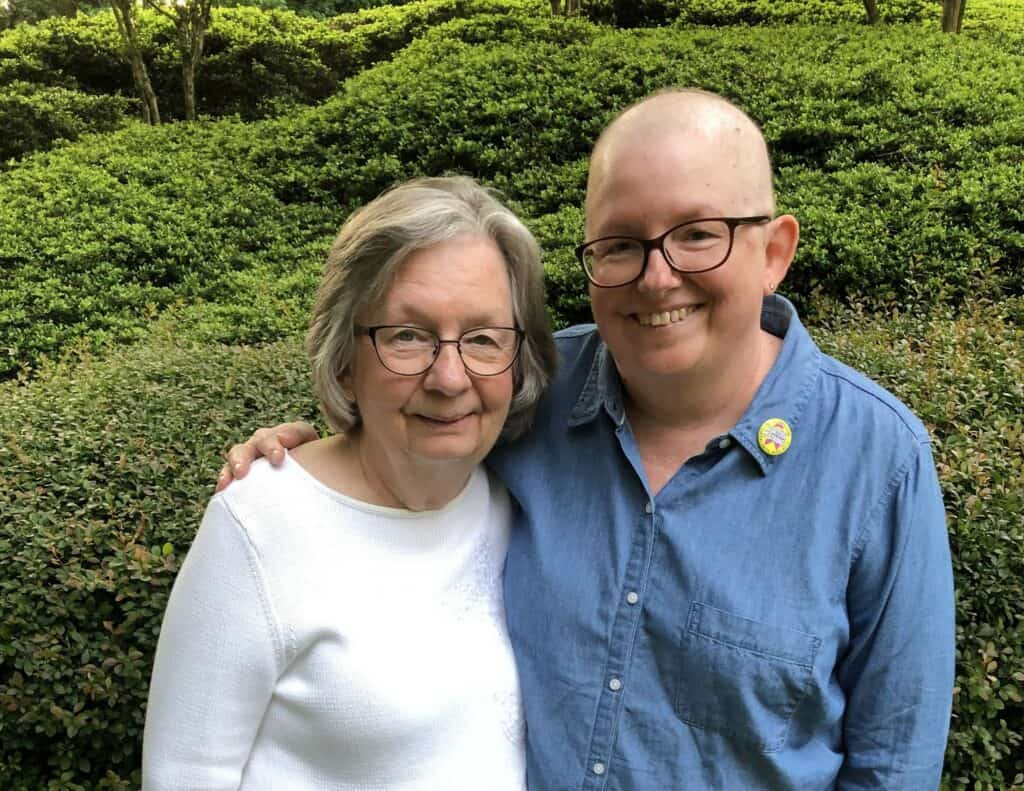 A breast cancer survivor during treatment, bald and wearing glasses, poses warmly with her mother. The two women stand close together, with arms around each other, in front of a green garden background. The daughter wears a blue shirt, and her mother is in a white top.
