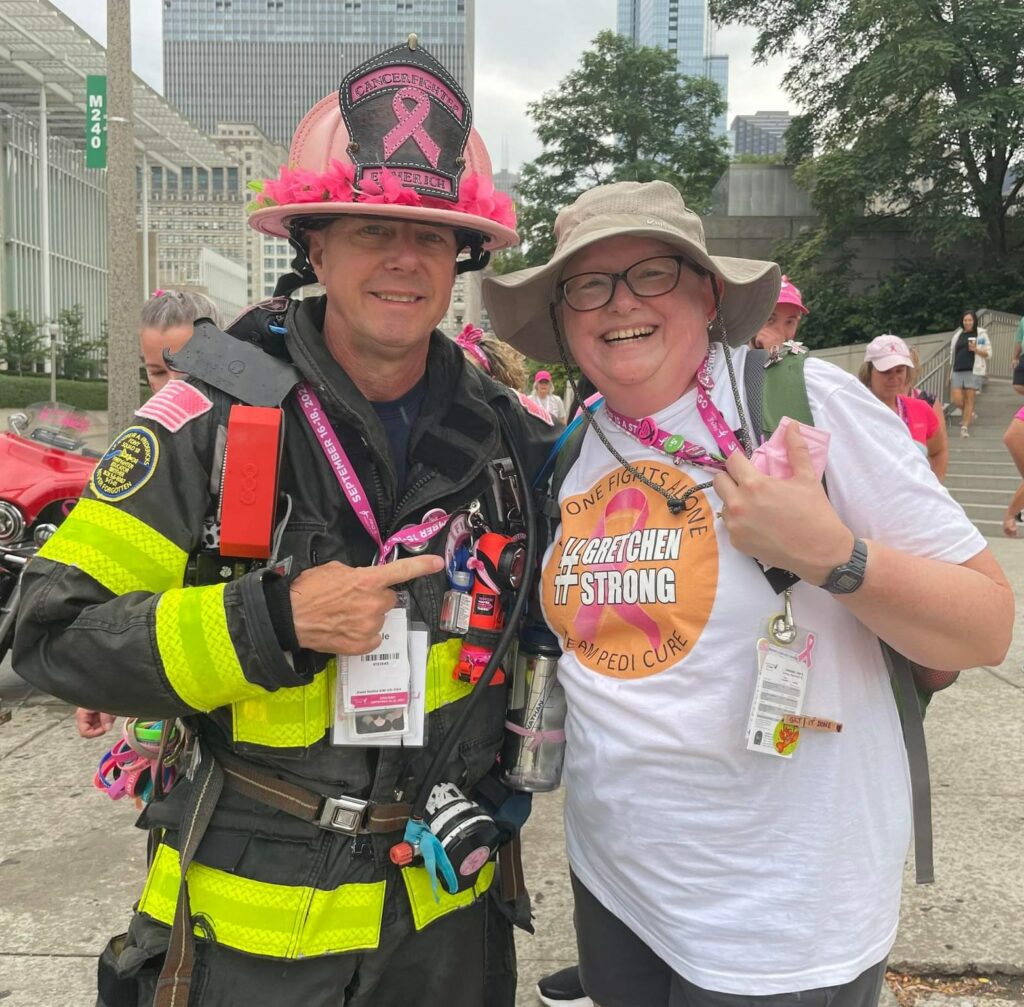 A firefighter in gear with a pink helmet at and gloves poses with a smiling woman wearing a white t-shirt that reads "Gretchen Strong" and a bucket hat. Both are wearing walkie-talkies and pink-themed accessories, standing outside in an urban setting at a Susan G. Komen 3-Day event. 