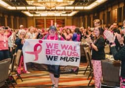 A smiling woman, wearing a Susan G. Komen hat and pink attire, proudly holds a banner that reads "We walk because we must" as she completes the Susan G. Komen 3-Day event. She is surrounded by a cheering crowd in a warmly lit ballroom, celebrating her as the final walker.