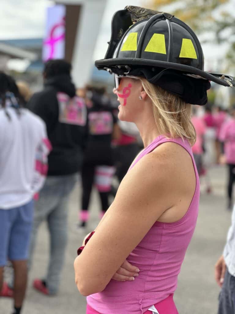 A woman wearing a firefighter helmet and a pink tank top stands with her arms crossed. She has a pink ribbon painted on her cheek, symbolizing breast cancer awareness. People in similar pink attire can be seen in the background, participating in an event.