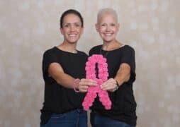 Two women standing side by side, both with short hair and wearing black shirts and jeans, smile at the camera. They hold a pink ribbon made of flowers, symbolizing breast cancer awareness. The background is softly blurred with light-colored bokeh.