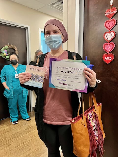 Jenn wears a mask. She's holding a certificate of completion for chemotherapy. She is smiling behind her mask. 