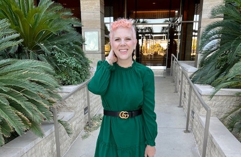 Jenn smiles at the camera. She's wearing a green dress and her hair, which is just starting to grow out after being bald, is colored pink.