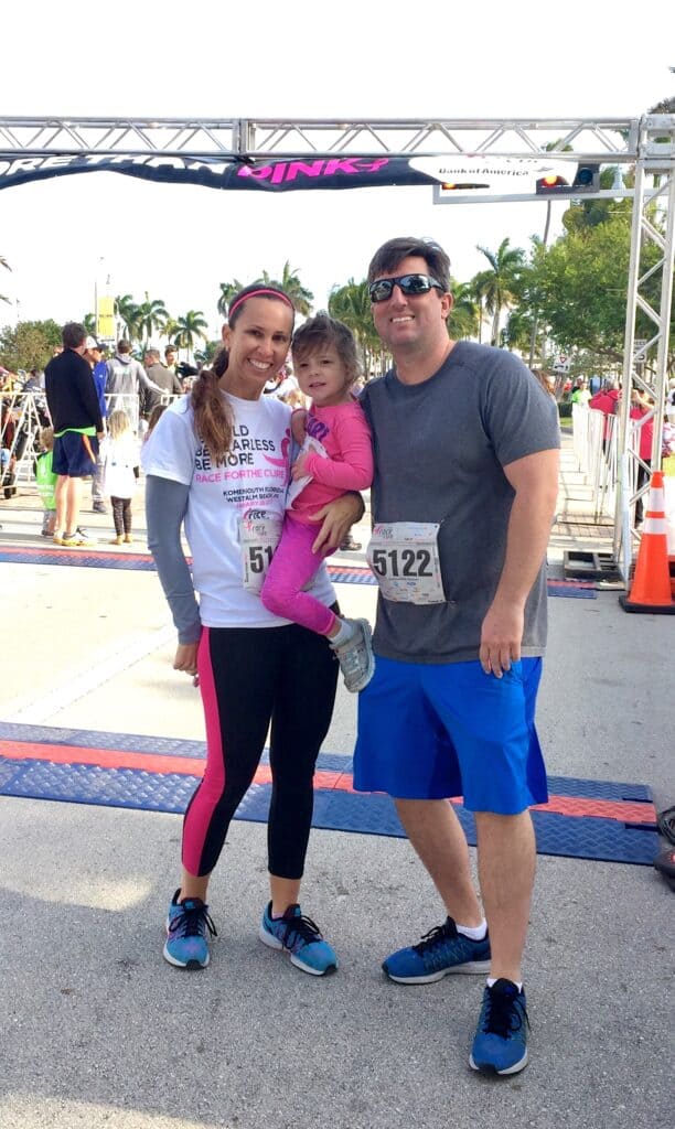 A woman, man, and young child stand together at a race finish line. The woman is holding the child, and both adults have race numbers pinned to their clothing. The background shows other participants and tropical trees.
