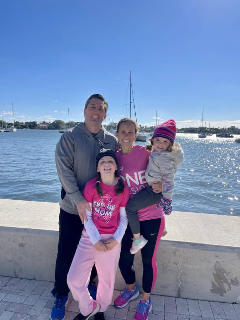 A family of four stands by a waterfront. The parents are smiling, with the father wearing a gray hoodie and the mother in a pink shirt holding the younger child. The children, one in pink and one in a colorful hat, smile under a clear blue sky.
