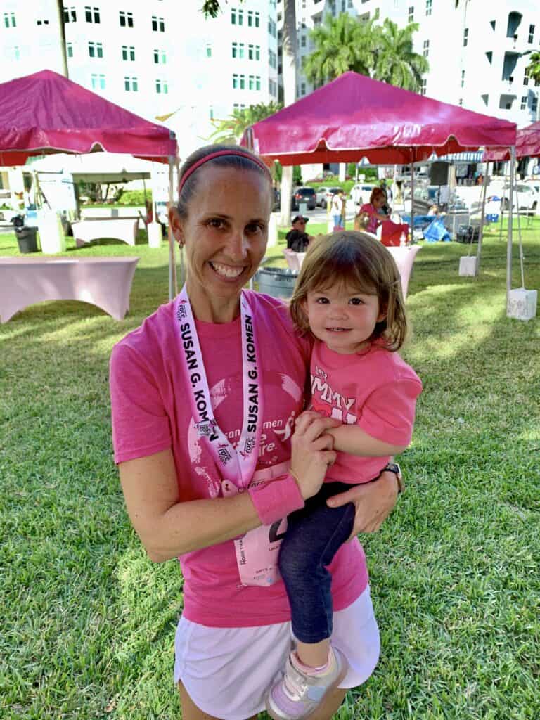 A woman wearing a pink shirt and medal stands on grass, holding a smiling child in her arms. They're in front of pink tents on a sunny day, at a Race for the Cure event.
