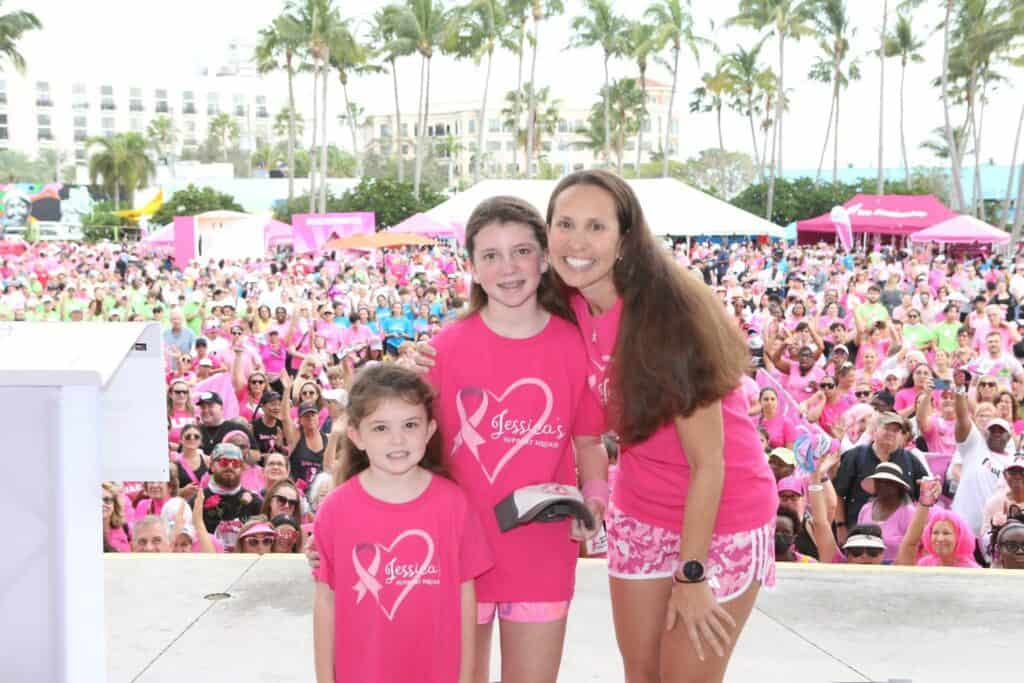 A woman and two children in pink shirts stand on a stage in front of a large crowd, all dressed in pink. The event appears festive, with palm trees and tents in the background on a sunny day.
