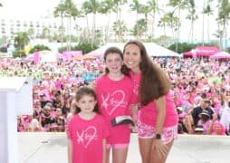 A woman and two children in pink shirts stand on a stage in front of a large crowd, all dressed in pink. The event appears festive, with palm trees and tents in the background on a sunny day.