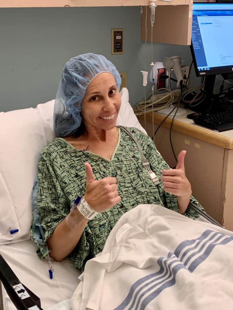 A patient in a hospital bed smiles and gives two thumbs up. They are wearing a hospital gown and blue surgical cap. Medical equipment and a computer are visible in the background.
