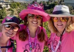 Three women dressed pink clothes and accessories are standing side by side smiling. There is a nice view of lush greenery behind them.