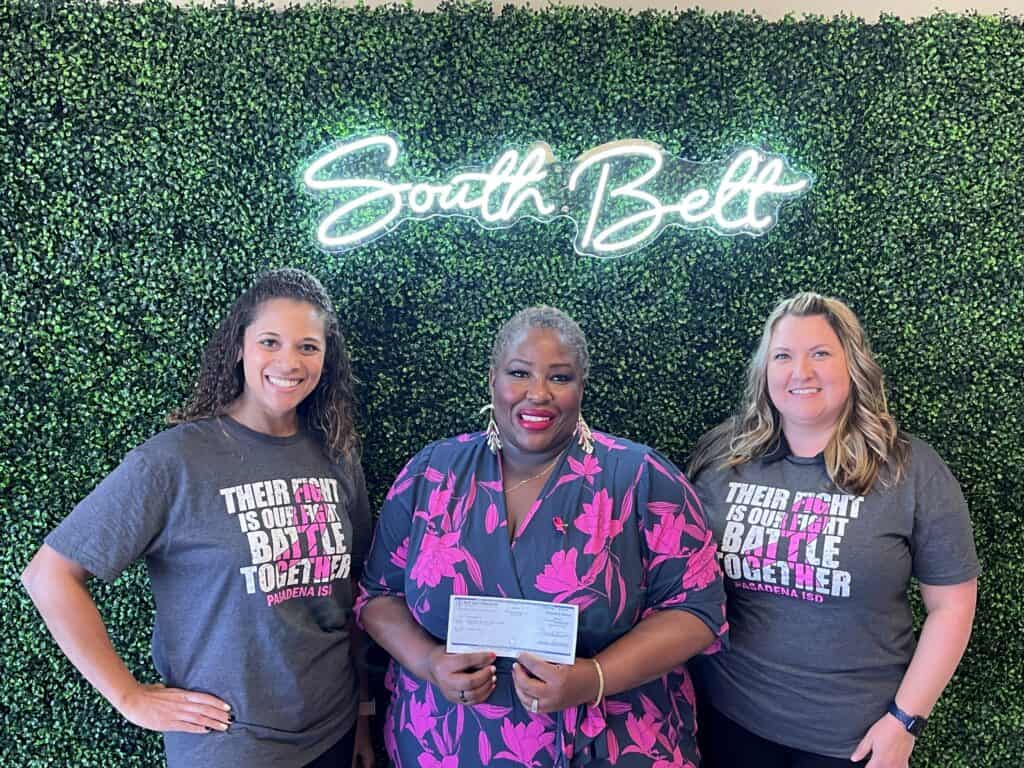 Three smiling women stand in front of a lush green wall with a neon sign that reads "South Belt." The woman in the center holds a check, while the others wear grey shirts with pink text saying, "Their Fight is Our Fight, Battle Together," supporting Susan G. Komen’s breast cancer awareness campaign.
