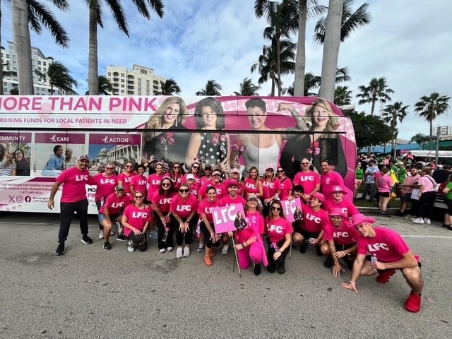 A large group of people wearing bright pink outfits and holding signs with 'LFC' gather in front of a decorated bus with 'More Than Pink' branding. The bus features images of breast cancer survivors and messaging about raising funds for local patients in need. The group is outdoors, surrounded by palm trees and a sunny sky, during what appears to be a breast cancer awareness walk or event.