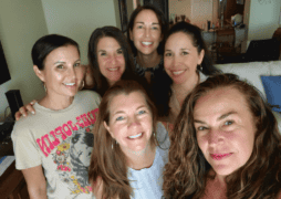 A group of six smiling women posing together indoors in a cozy living room. The women are gathered closely, showing camaraderie and joy, with a mix of casual and stylish outfits. Behind them, the room features a sofa with blue pillows and a dining area with warm lighting.