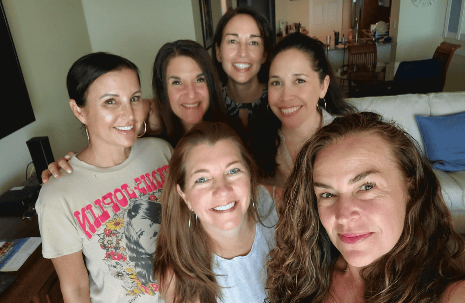 A group of six smiling women posing together indoors in a cozy living room. The women are gathered closely, showing camaraderie and joy, with a mix of casual and stylish outfits. Behind them, the room features a sofa with blue pillows and a dining area with warm lighting.