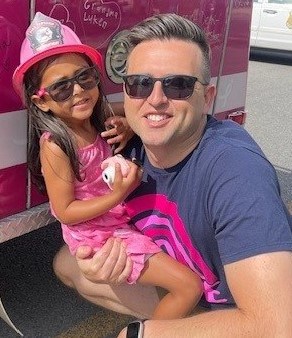 A man wearing sunglasses and a dark t-shirt kneels beside a young girl in a pink dress and toy firefighter hat. They are both smiling. The girl is holding a small white toy and they are in front of a pink emergency vehicle decorated with drawings and text.