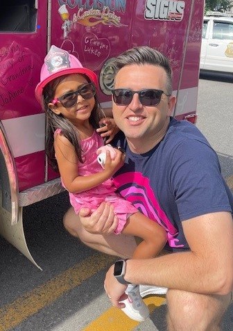 A man wearing sunglasses and a dark t-shirt kneels beside a young girl in a pink dress and toy firefighter hat. They are both smiling. The girl is holding a small white toy and they are in front of a pink emergency vehicle decorated with drawings and text.
