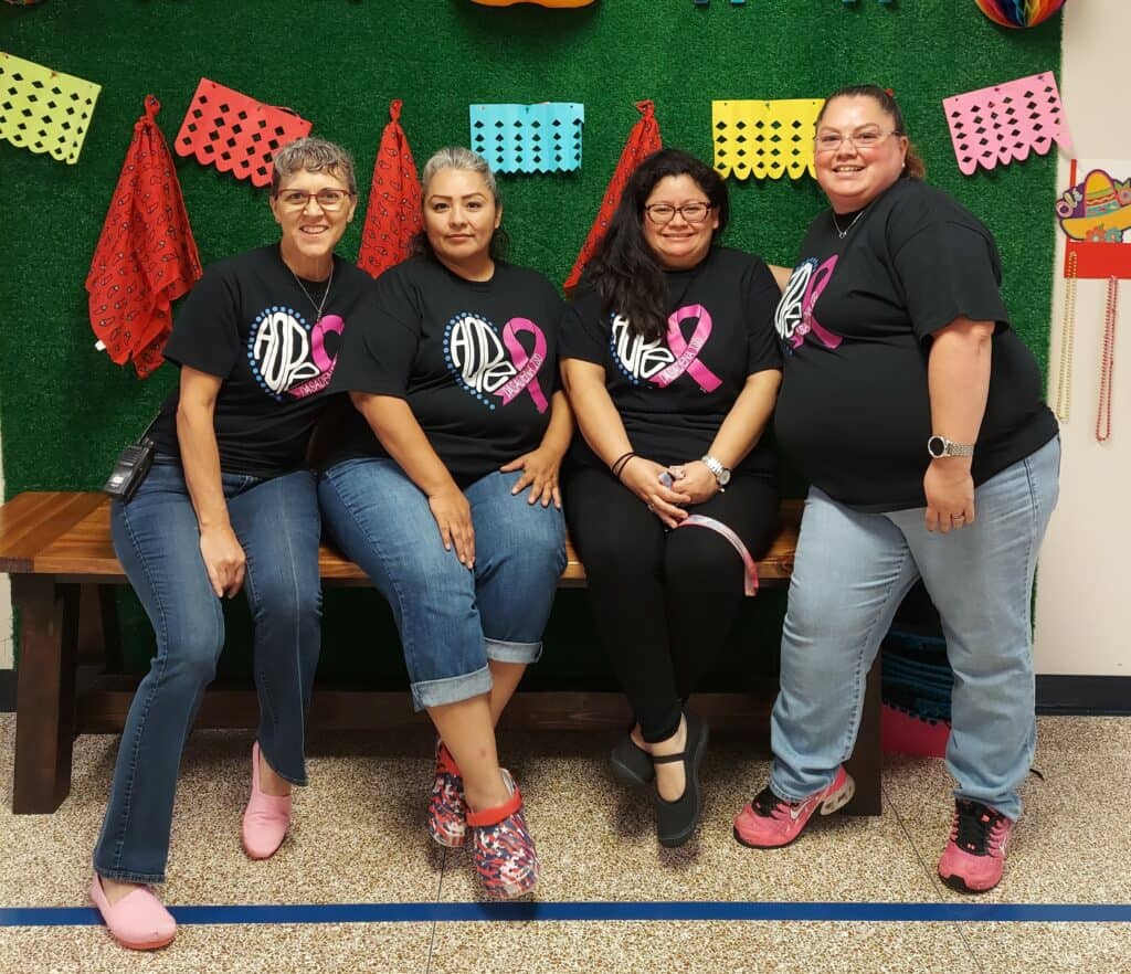 Four people are sitting on a bench in front of a green wall with colorful decorations. They are smiling and wearing matching black T-shirts with a pink ribbon design, showing their support for Go Pink for the Cure. The setting appears to be festive.