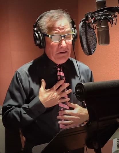 A man with glasses is in a recording studio, wearing headphones. Donning a pink tie, he stands before a microphone, his hand to his chest as he reads from a script on a music stand. The backdrop of soundproofing panels attests to the importance of his message for the breast cancer ambassador fundraising campaign.
