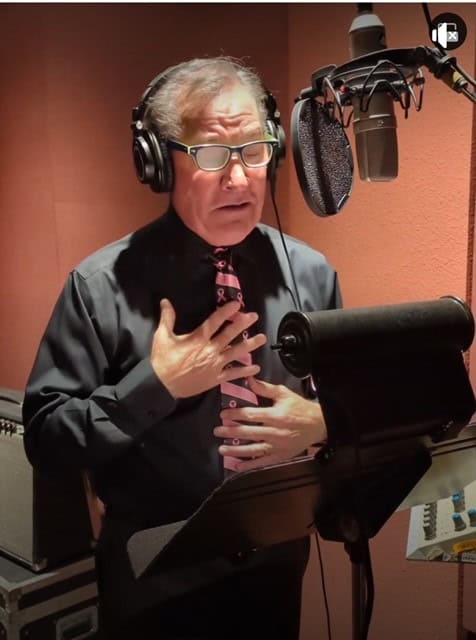 A man with glasses is in a recording studio, wearing headphones. Donning a pink tie, he stands before a microphone, his hand to his chest as he reads from a script on a music stand. The backdrop of soundproofing panels attests to the importance of his message for the breast cancer fundraising campaign.
