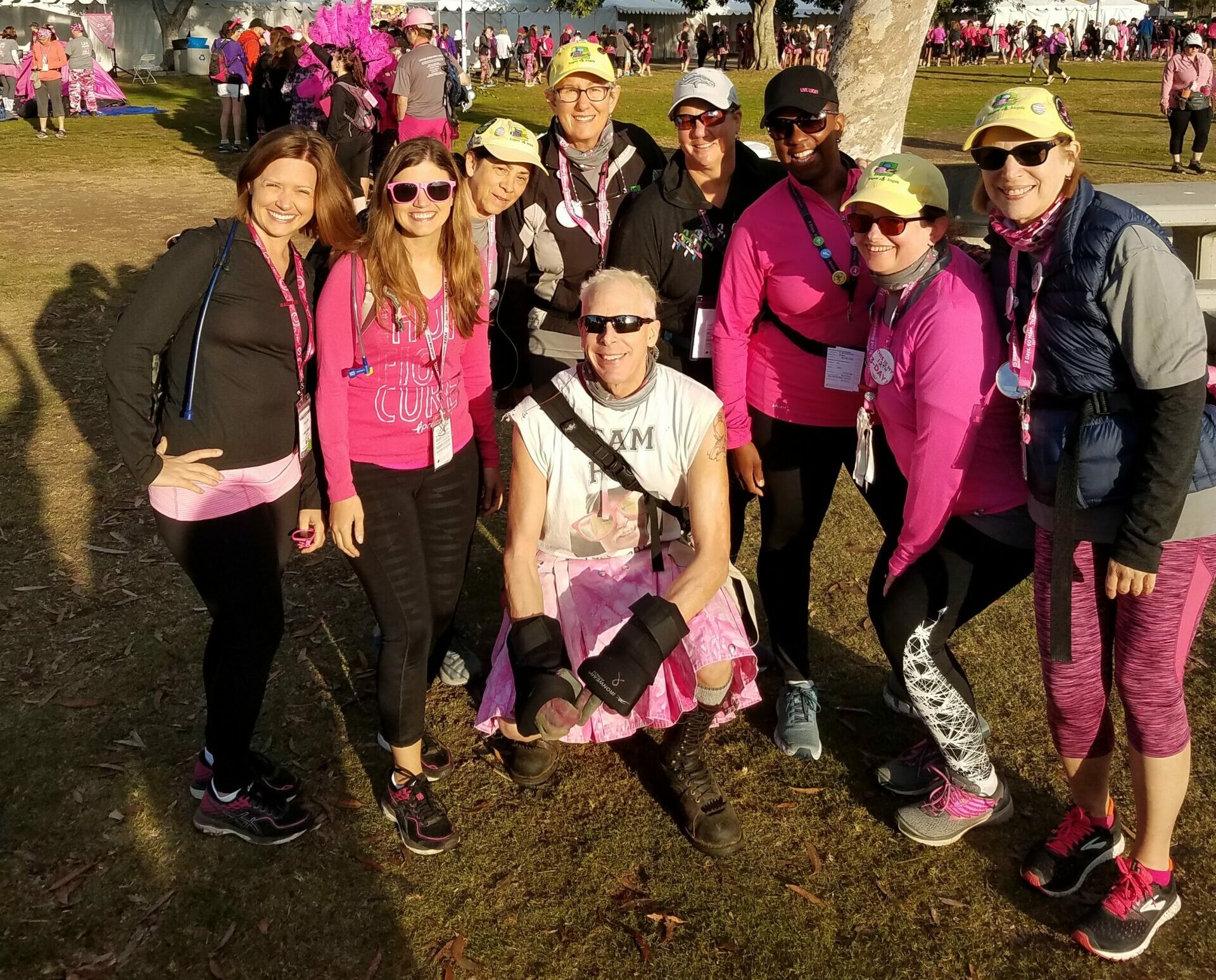 A group of people, mostly women, pose together outdoors at an event. They are wearing matching pink outfits and accessories, indicating participation in a Susan G. Komen 3-Day breast cancer awareness walk. Trees and tents can be seen in the background under a partly cloudy sky.
