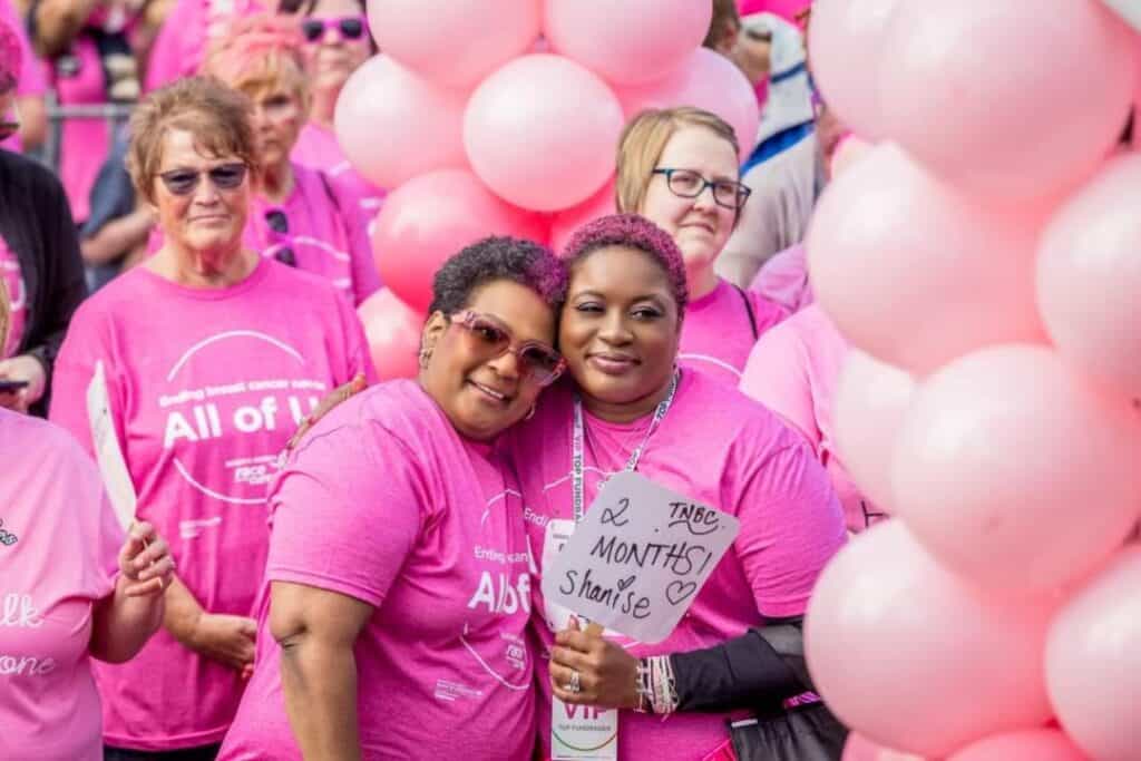 Shanise and a loved one wear pink shirts, surrounded by people in pink and pink balloons.