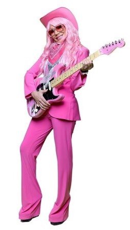 Person dressed in a bright pink outfit, including a suit, cowboy hat, and bandana, holding a pink electric guitar. They have long, wavy hair and are also wearing sunglasses. The background is plain white. This vibrant look is part of a Susan G. Komen breast cancer fundraising campaign.