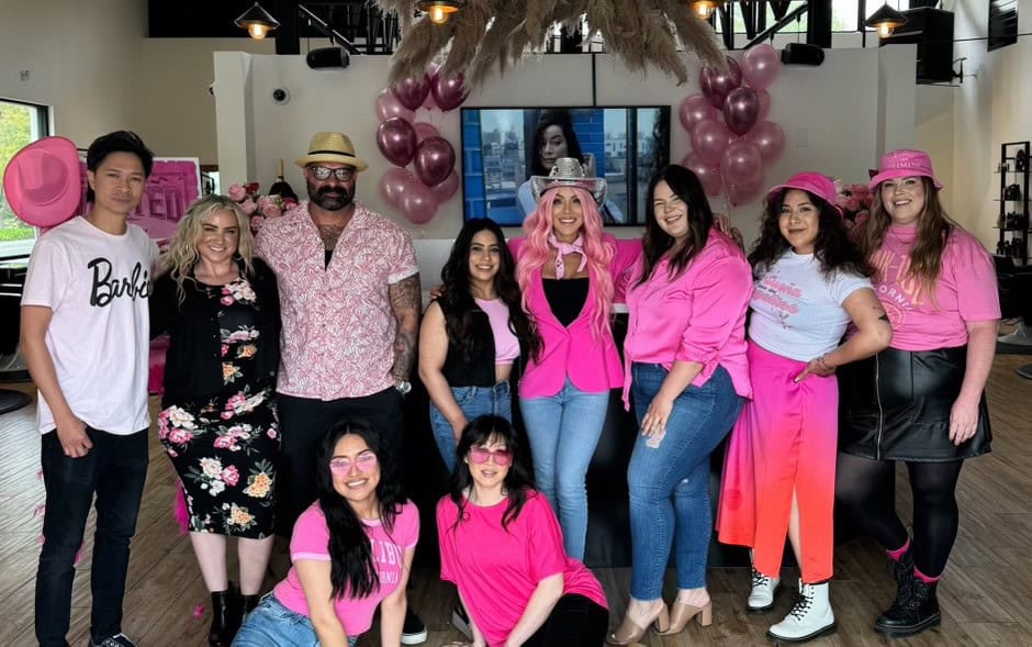 A group of people posing indoors, all dressed in various shades of pink in festive attire for a Susan G. Komen breast cancer fundraising event. Some are wearing hats and sunglasses, and there are pink balloons in the background. A large television screen displays an image, and the setting appears to be a modern event space.
