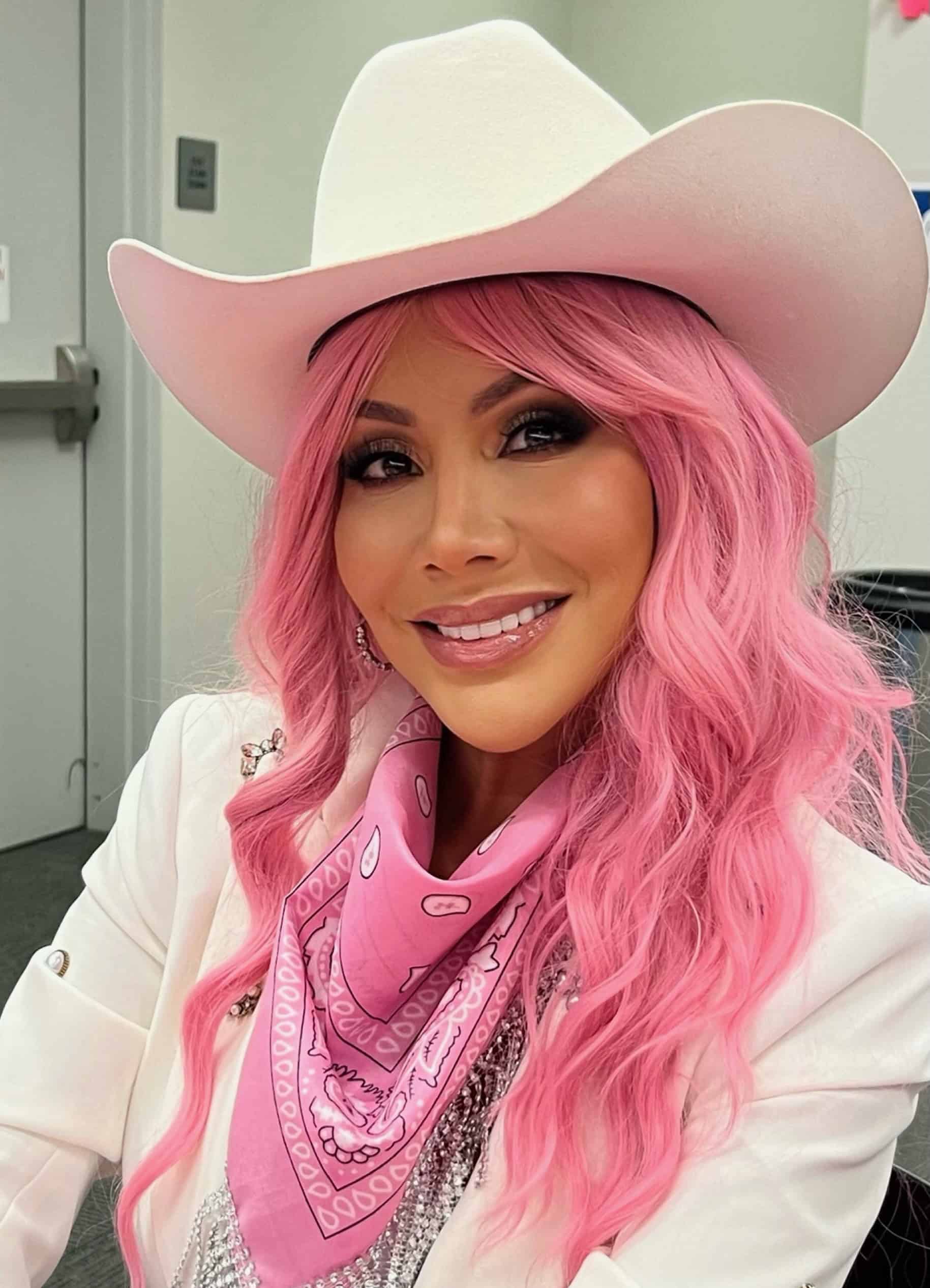 A woman wearing a white jacket with a sparkly pink bandana, bright pink wig and white cowboy hat. She smiles at the camera.
