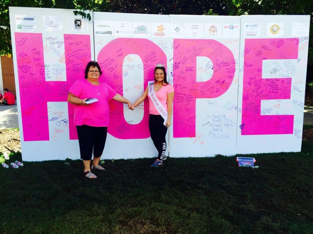 Two people in pink stand in front of a large "HOPE" sign covered in handwritten messages.