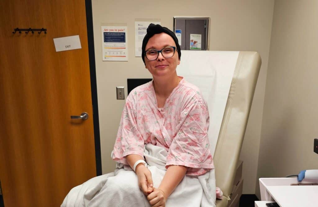 Sierra is in an exam room. She is wearing a robe and has a scarf on her head. She is smiling.